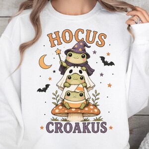 May include: A white sweatshirt featuring a graphic of three stacked frogs on a mushroom. The top frog wears a purple wizard hat and cape, the middle is under a ghost sheet, and the bottom wears a candy corn hat. Text reads HOCUS CROAKUS.