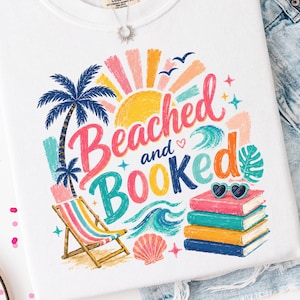Op de afbeelding: Een wit t-shirt met een kleurrijke afbeelding en de tekst Beached and Booked. Het ontwerp bevat een gele zon, blauwe palmbomen, een gestreepte strandstoel, een roze schelp, oceaangolven en een stapel van vier boeken met een hartvormige zonnebril erop.