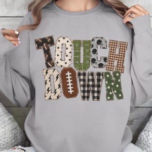 May include: A light gray crewneck sweatshirt featuring the word TOUCHDOWN in large block letters. Each letter has a unique pattern including footballs, stars, gridiron field markings, helmets, plaid, lightning bolts, and a football lace design.