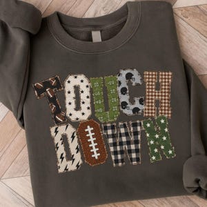 May include: A charcoal gray crewneck sweatshirt featuring the word TOUCHDOWN in large, multi-patterned block letters. The letters include designs like footballs, stars, field markings, helmets, plaid, lightning bolts, and a brown football shape.