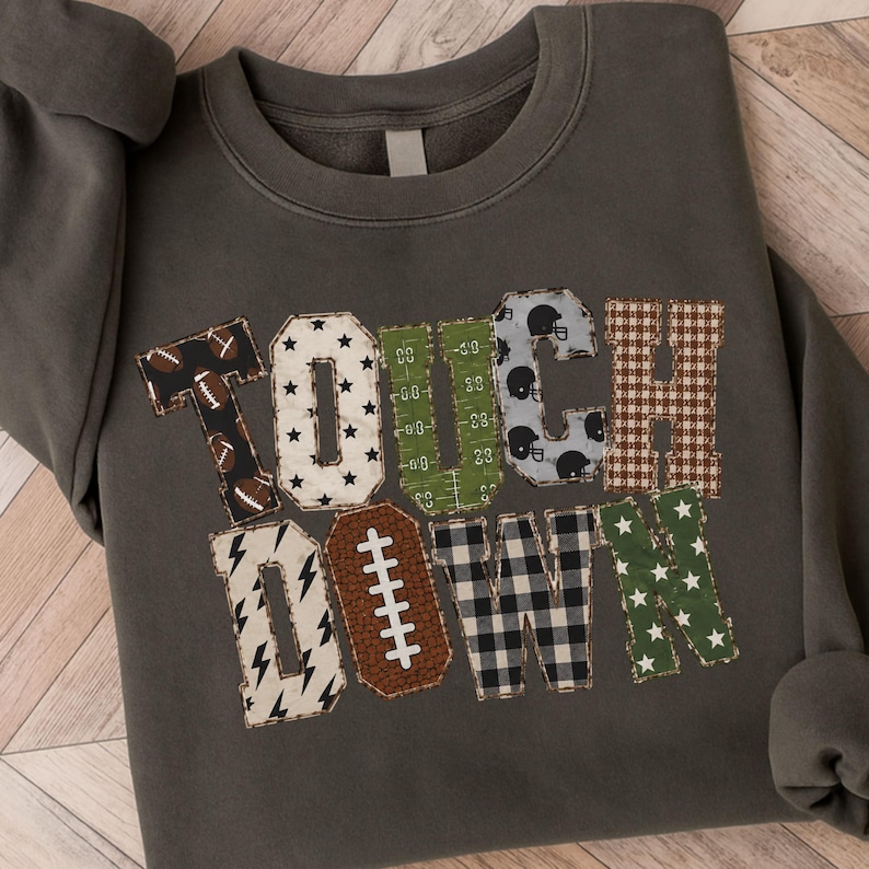 May include: A charcoal gray crewneck sweatshirt featuring the word TOUCHDOWN in large, multi-patterned block letters. The letters include designs like footballs, stars, field markings, helmets, plaid, lightning bolts, and a brown football shape.