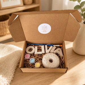 Könnte beinhalten: Ein Bastelset in einer offenen Pappschachtel. Das Set enthält den Namen "OLIVIA" in einzelnen Buchstaben, einen donut-förmigen Gegenstand, Farbtöpfe und einen Pinsel. Ein Aufkleber auf der Box lautet "Let's make something magical."
