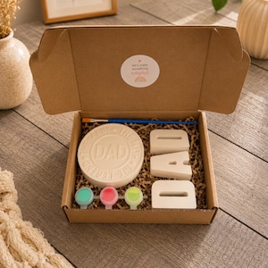 May include: A craft kit in a cardboard box, containing a round ceramic piece with "DAD" text, individual letter cutouts spelling "DAD", paint pots, and a paintbrush. The box has a sticker that says "let's make something magical".