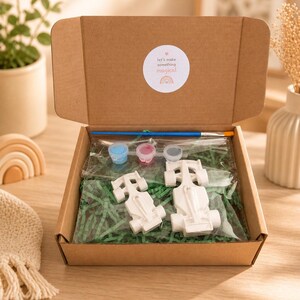 Könnte beinhalten: Geöffnete Pappschachtel mit einem Mal-dein-eigenes-Rennwagen-Set. Das Set enthält zwei weiße Rennwagenformen, Farbtöpfe in Blau, Rosa und Grau, einen Pinsel und grüne Papierfüllung. Die Schachtel hat einen Aufkleber mit der Aufschrift "let's make something magical".