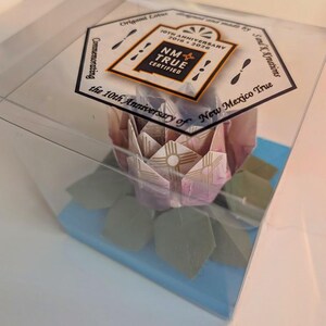 May include: A clear plastic box holds a paper origami lotus flower with pink and white petals and green leaves. The box sits on a blue base and has a label reading "NM True Certified" and "10th Anniversary 2016-2026".