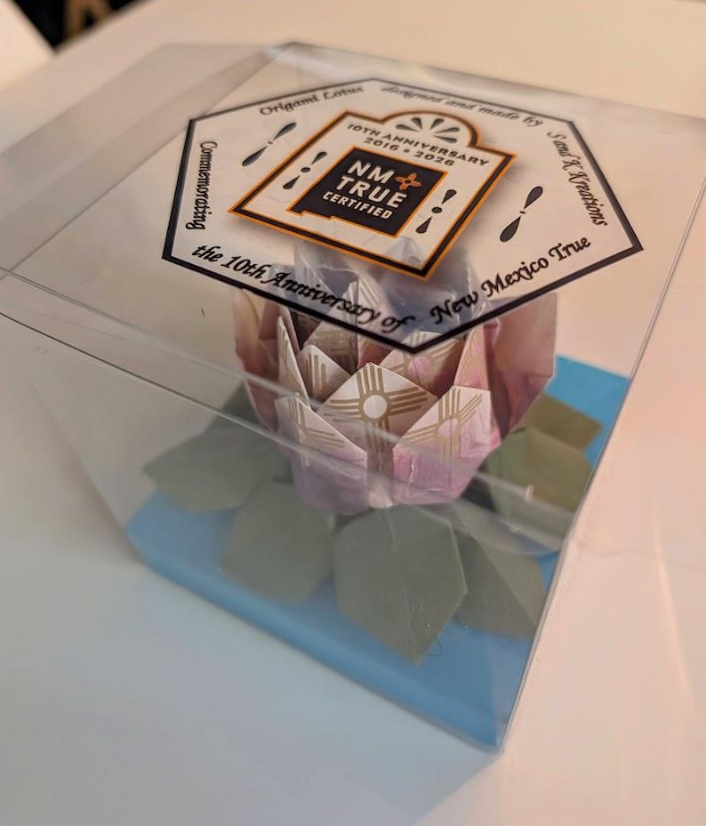 May include: A clear plastic box holds a paper origami lotus flower with pink and white petals and green leaves. The box sits on a blue base and has a label reading "NM True Certified" and "10th Anniversary 2016-2026".