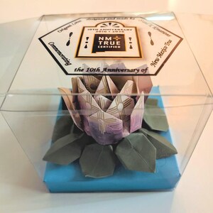 May include: An origami lotus flower, presented in a clear plastic box. The flower is crafted from purple and white paper, with green leaves. The box sits on a blue base and features a label with "NM True Certified" and "10th Anniversary".