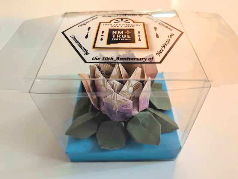 May include: An origami lotus flower, presented in a clear plastic box. The flower is crafted from purple and white paper, with green leaves. The box sits on a blue base and features a label with "NM True Certified" and "10th Anniversary".