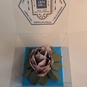 May include: An origami rose made from paper, displayed in a clear box. The rose is a mix of pink and purple hues, with green paper leaves. The box sits on a blue base, and the top of the box has text.