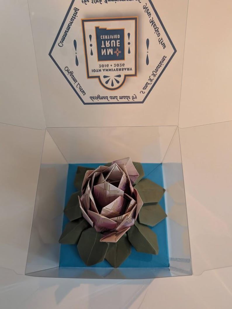 May include: An origami rose made from paper, displayed in a clear box. The rose is a mix of pink and purple hues, with green paper leaves. The box sits on a blue base, and the top of the box has text.