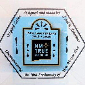 May include: A hexagonal emblem with a light blue background, featuring text that reads "10TH ANNIVERSARY 2016 &bull; 2026" and "NM TRUE CERTIFIED." The emblem is surrounded by text that says "Origami Lotus" and "New Mexico True."