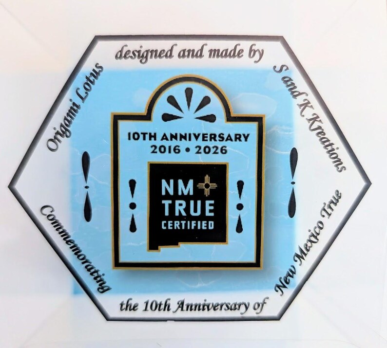 May include: A hexagonal emblem with a light blue background, featuring text that reads "10TH ANNIVERSARY 2016 &bull; 2026" and "NM TRUE CERTIFIED." The emblem is surrounded by text that says "Origami Lotus" and "New Mexico True."