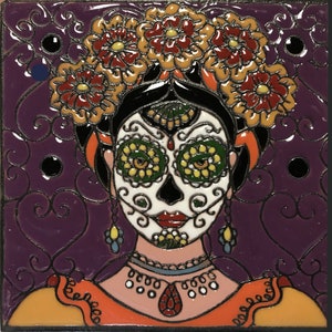 May include: A colorful ceramic tile featuring a Day of the Dead sugar skull with a floral crown. The skull is painted in white with black outlines and green and yellow eyes. The background is a dark purple with a swirling pattern.
