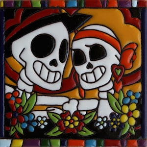 May include: A colorful ceramic tile featuring two skeletons with smiling faces, wearing hats and flowers. The tile has a black background with a border of colorful stripes.