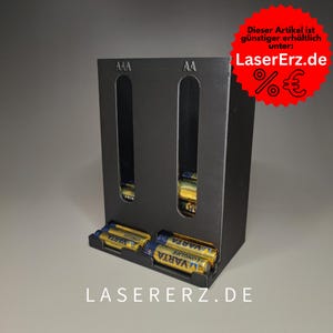 Könnte beinhalten: Schwarzer Batterie-Organizer mit zwei Fächern, die mit "AAA" und "AA" beschriftet sind. Der Organizer hat einen Schlitz für Batterien, die unten eingesetzt werden können. Der Organizer ist aus schwarzem Acryl und hat ein weißes Etikett auf der Unterseite, auf dem "LASERERZ.DE" steht.