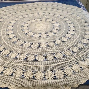 May include: A cream-coloured, circular crocheted tablecloth with an intricate openwork design. The pattern features concentric circles with floral motifs and a scalloped edge. The tablecloth is displayed on a blue bedspread.