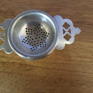 May include: A silver tea strainer with a round bowl and decorative handles. The bowl has a pattern of small holes in the shape of a star. The handles have a clover-like design. The strainer is made of metal and is designed for steeping loose leaf tea.