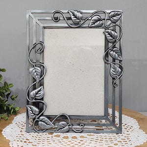 May include: A rectangular silver-colored picture frame with a decorative leaf and vine design. The frame has a vertical orientation and a neutral-colored backing. The leaves are detailed and add a natural element to the frame's design.