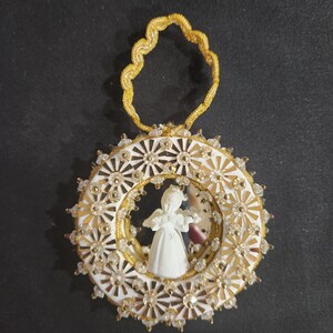 May include: A white and gold Christmas ornament featuring a central figure of a person in a dress. The ornament has a circular design with a white background, gold accents, and clear crystal-like embellishments. A gold loop is at the top.