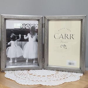 May include: A hinged silver-colored photo frame with a decorative border. One side displays a black and white photo of two young girls in white dresses. The other side features the text "CARR Frames" and a decorative emblem.