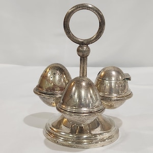 May include: A silver-colored egg-shaped condiment set with a circular handle. The set includes a salt shaker with small holes, a pepper shaker, and a lidded container, all resting on a round base. The design is vintage and elegant.