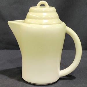 May include: A cream-colored ceramic teapot with a lid that has a ridged design. The teapot has a rounded body and a curved handle.