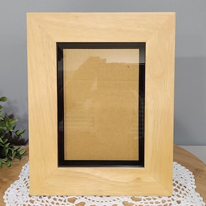 May include: A light-colored wooden picture frame with a black inner border. The frame is rectangular and has a simple, clean design. The frame is empty, with a cardboard backing visible.