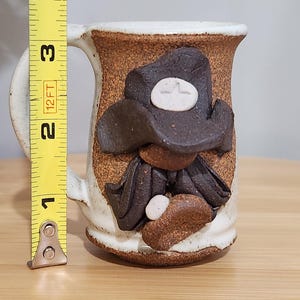May include: Handmade ceramic mug with a cowboy design. The mug is approximately 3 inches tall and features a brown hat, scarf, and other details. The mug has a white handle and rim, and a textured brown body.