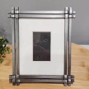 May include: A rectangular metal picture frame with a dark gray finish. The frame has a white mat and a small, dark image. The frame is on a wooden surface with green foliage in the background.