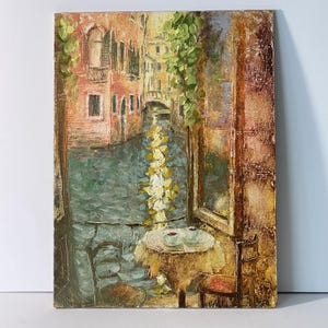 Può includere: Dipinto a olio di una scena del canale veneziano. L'opera raffigura un canale con edifici su entrambi i lati, un ponte in lontananza e un tavolino con due tazze. La tavolozza dei colori comprende sfumature di marrone, verde e blu.