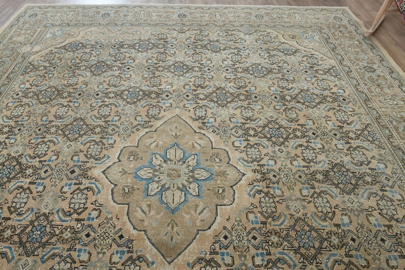 May include: A large, rectangular area rug with an intricate, traditional design. The rug features a central medallion with a floral motif in shades of blue and beige, surrounded by a repeating pattern in similar tones. The overall color palette is muted, with a vintage aesthetic.