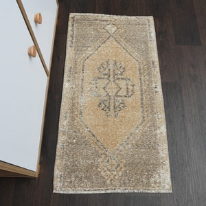 May include: A beige and brown vintage rug with a geometric pattern in the center. The rug has a frayed edge and is laying on a dark wood floor.