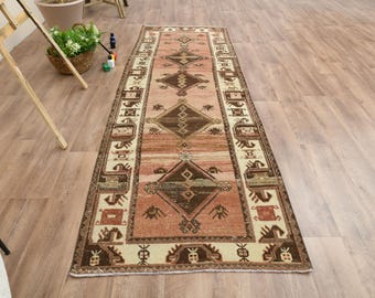 Puede incluir: Una alfombra larga y rectangular con un diseño persa tradicional. La alfombra tiene un fondo crema con un patrón geométrico marrón y rosa. La alfombra es perfecta para agregar un toque de estilo tradicional a cualquier habitación.