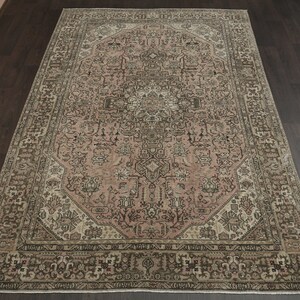 May include: An ornate, rectangular rug with a faded floral design. The rug has a central medallion in pink and brown hues, framed by a border in similar tones. It is displayed on a dark wooden floor, showcasing its intricate details.
