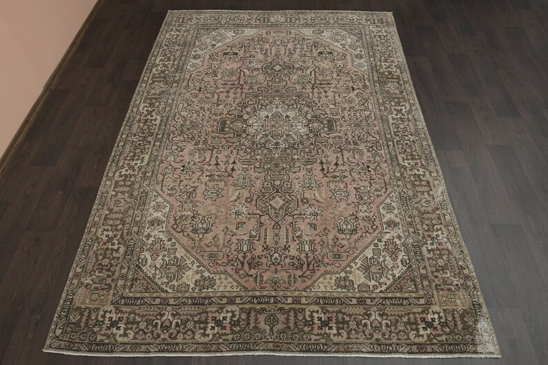 May include: An ornate, rectangular rug with a faded floral design. The rug has a central medallion in pink and brown hues, framed by a border in similar tones. It is displayed on a dark wooden floor, showcasing its intricate details.