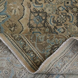 May include: An antique rug with a distressed, vintage look. The rug features a complex pattern in shades of brown, beige, and blue. The edges are frayed, adding to the rug's aged appearance. The rug is folded over, revealing the underside.