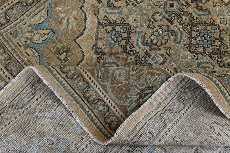 May include: An antique rug with a distressed, vintage look. The rug features a complex pattern in shades of brown, beige, and blue. The edges are frayed, adding to the rug's aged appearance. The rug is folded over, revealing the underside.