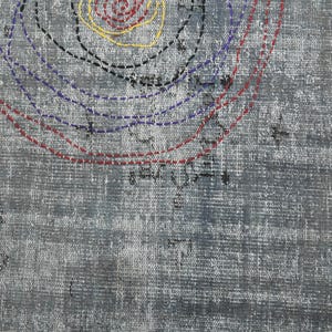 May include: A close-up of a gray textured fabric with a circular design in red, blue, black, and yellow thread. The design is made up of concentric circles with dashed lines.