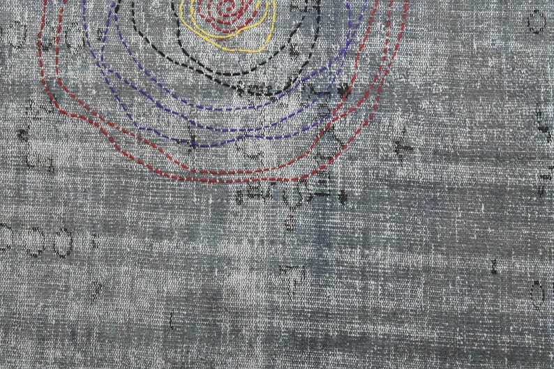 May include: A close-up of a gray textured fabric with a circular design in red, blue, black, and yellow thread. The design is made up of concentric circles with dashed lines.