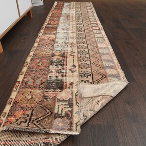 Pode incluir: Um tapete corredor vintage com um esquema de cores marrom, bege e rosa. O tapete tem um padrão geométrico com detalhes intrincados. O tapete está dobrado ao meio e colocado sobre um piso de madeira escura.