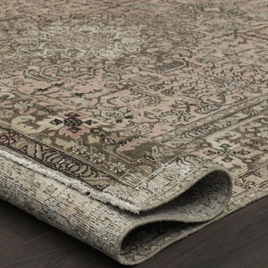 May include: A rolled-up, vintage rug with a faded, distressed look. It has a complex floral pattern in beige, brown, and pink. The edges are frayed, adding to its antique charm. The rug is on a dark wood surface.