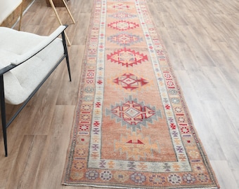 Puede incluir: Una alfombra de pasillo larga y estrecha con un color base naranja descolorido y una serie de patrones geométricos de diamantes repetidos en tonos rojo, azul y crema. La alfombra tiene un borde decorativo con pequeños motivos.