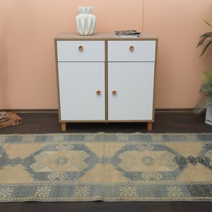 May include: A light blue and beige patterned rug with a geometric design. The rug is laid out on a wooden floor in front of a white cabinet with two drawers and two doors. A potted plant is on the right side of the image.