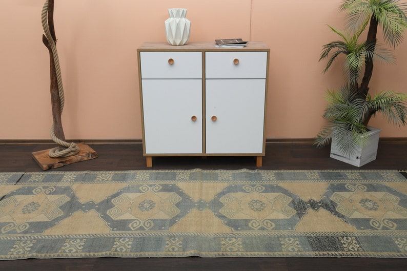 May include: A light blue and beige patterned rug with a geometric design. The rug is laid out on a wooden floor in front of a white cabinet with two drawers and two doors. A potted plant is on the right side of the image.