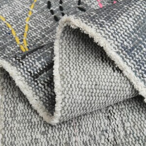 May include: Close-up of a gray and white woven rug with a fringe edge. The rug has a geometric pattern with black, yellow, and pink accents.