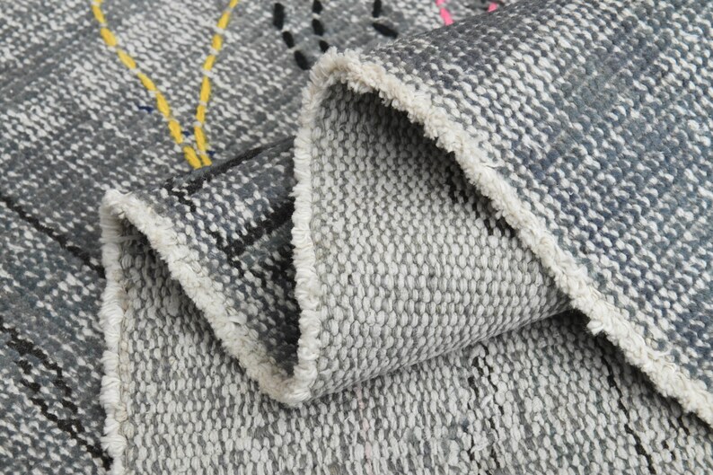 May include: Close-up of a gray and white woven rug with a fringe edge. The rug has a geometric pattern with black, yellow, and pink accents.