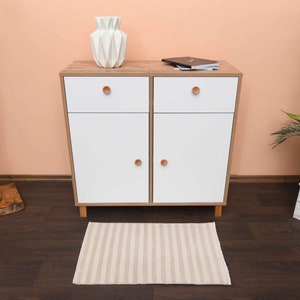 May include: A white cabinet with two drawers and two doors. The cabinet has wooden legs and wooden handles. The cabinet is sitting on a striped rug in front of a peach wall.