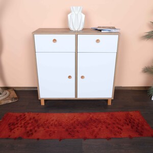 May include: A red runner rug with a textured pattern is placed on a dark wood floor. A white cabinet with wooden knobs and a decorative white vase sit against a peach-colored wall. A decorative tree trunk with rope and a potted plant are also in the scene.