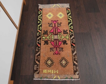 May include: A brown and beige runner rug with a geometric pattern in shades of pink, yellow, and black. The rug has a fringe border and the text "HEMEIY" is woven into the bottom of the rug.