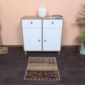 Può includere: Un piccolo tappeto rettangolare con un design patchwork con vari colori e motivi. Il tappeto è posto su un pavimento in legno scuro davanti a un mobile bianco con pomelli in legno. Un tronco d'albero decorativo con corda e una pianta in vaso sono anche nell'immagine.